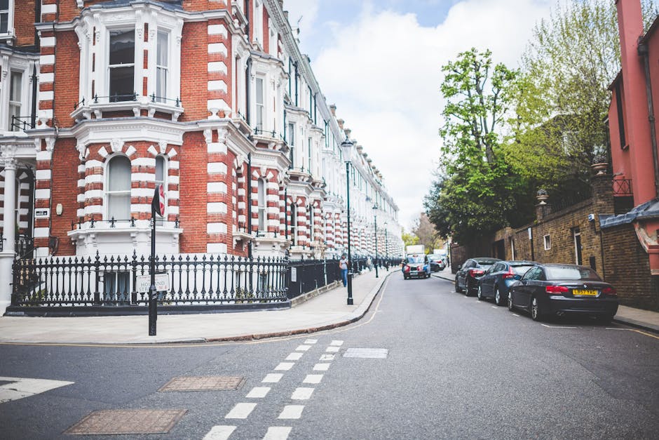 Image depicting a street scene in Wandle Park area with residential Victorian-style terraced houses featuring red and white brick facades, bay windows, and decorative architectural details. Parked cars line the pavement on the right side, including a black car closest to the foreground and additional vehicles further along. A black wrought iron fence surrounds a small front garden space in front of one of the houses. The pavement is clean and well-maintained, with a few pedestrians visible in the background. The sky is partly cloudy with patches of blue, and trees with green foliage provide some natural shade. This scene illustrates the typical environment where house removals and furniture transport activities may take place, aligning with the services offered by Removals Colliers Wood during home relocation or packing and moving processes.