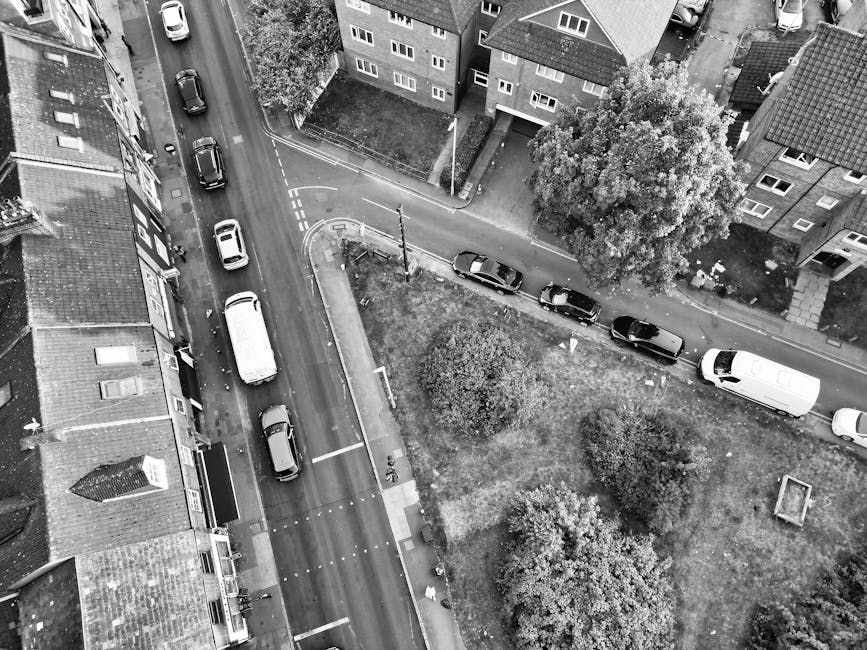 An aerial black-and-white photograph capturing a residential street scene during a home relocation process. The image shows a row of parked cars along a narrow road adjacent to a small grassy area with three trees. A white van with a boxy shape is parked facing the curb, indicating it is being used for furniture transport or packing and moving activities. Several large cardboard boxes and wrapped furniture items are visible near the van and on the pavement, suggesting recent packing or loading. Part of a mobile trolley or hand truck can be seen near the boxes, used for lifting or carrying heavy items during the furniture transport. The scene is illuminated by natural daylight, with shadows cast by the trees and buildings. The surrounding residential buildings are multi-storey, with flat roofs and multiple windows, typical of urban housing areas. This setting appears to be part of a property clearance or home relocation arranged by Removals Colliers Wood, with vehicles and furniture positioned for efficient loadings and transport to a new residence.