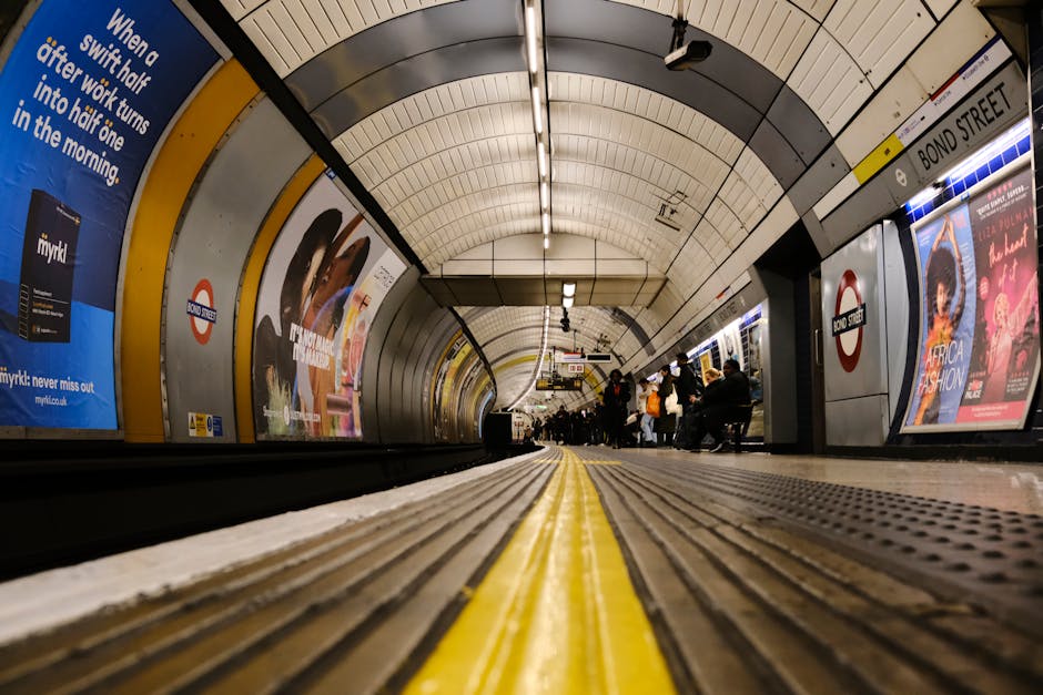 A view from the ground level inside London Underground's Bond Street station, showing the platform with a yellow safety line in the foreground, metal railings along the edges, and advertising posters on the curved white tiled walls. Passengers are seated on benches and waiting for trains, with some standing and holding luggage or bags. Overhead, fluorescent lighting illuminates the station, and an arching ceiling with metal panels extends overhead. The scene reflects a typical station environment used during home relocation or furniture transport, where moving companies like Removals Colliers Wood may coordinate loading or transferring belongings onto trains or into transport vans outside the station entrance.