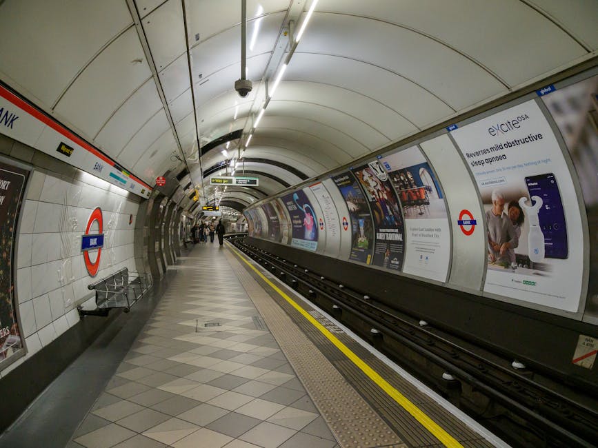 The image depicts a section of the London Underground station at Colliers Wood, featuring a curved platform with a tiled floor and white tiled walls. On the left side, there is a roundel sign with the station name 'MERTON PARK' visible, alongside a metal bench nearby. The platform has a yellow line along the edge, indicating safety boundaries, with a tactile paving strip for visually impaired passengers. Overhead, curved lighting fixtures provide illumination, and a security camera is mounted on the ceiling. Several passengers are seen standing or walking along the platform in the distance. The advertisement walls along the right side display various posters, including one promoting sleep aid devices. The scene captures a clean, well-maintained underground station environment, emphasizing the typical setting for London commuters, with visible elements related to transport logistics and station infrastructure, in line with [COMPANY_NAME]'s focus on assisting with house removals and home relocation services near Colliers Wood.