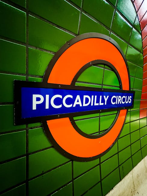 A view from the ground level inside London Underground's Bond Street station, showing the platform with a yellow safety line in the foreground, metal railings along the edges, and advertising posters on the curved white tiled walls. Passengers are seated on benches and waiting for trains, with some standing and holding luggage or bags. Overhead, fluorescent lighting illuminates the station, and an arching ceiling with metal panels extends overhead. The scene reflects a typical station environment used during home relocation or furniture transport, where moving companies like Removals Colliers Wood may coordinate loading or transferring belongings onto trains or into transport vans outside the station entrance.